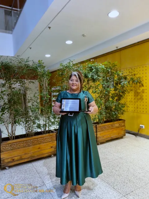 Sol Lima de vestido verde segura a placa de premiação no espaço da cerimônia.