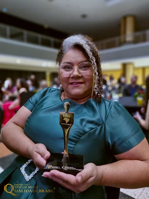 Sol Lima sentada na cerimônia, segurando o troféu Top of Mind Qualidade Brasil.