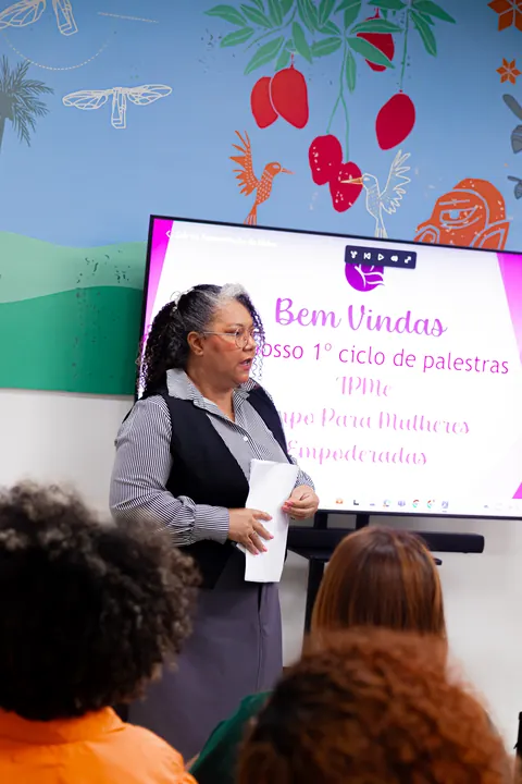 Mulher fala ao microfone diante de uma tela de boas-vindas.